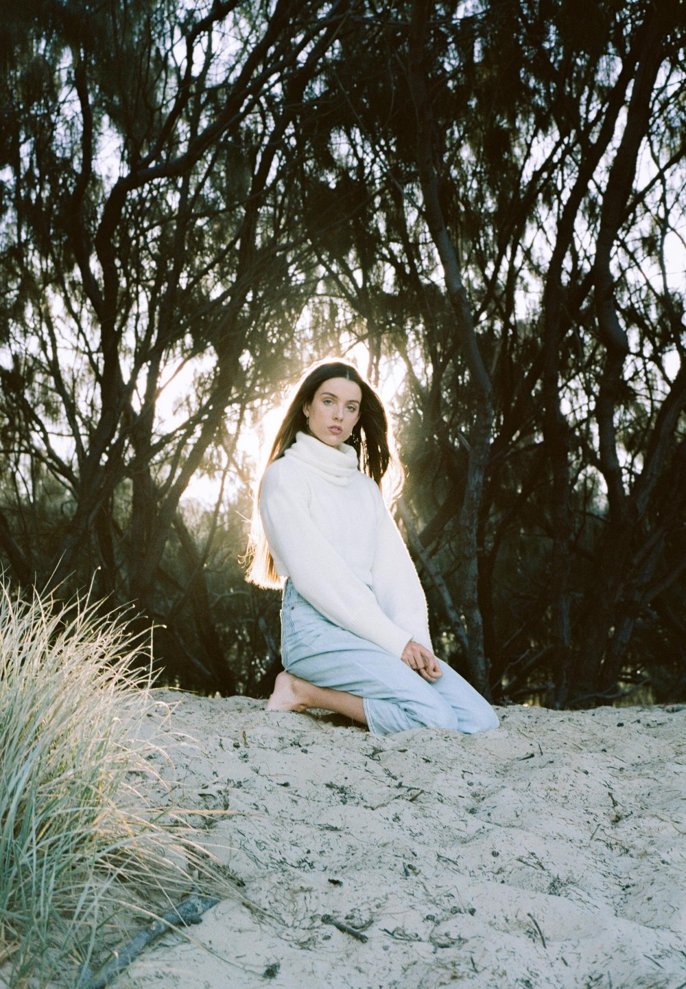 A woman kneels on a sandy dune with trees and bright light behind her.