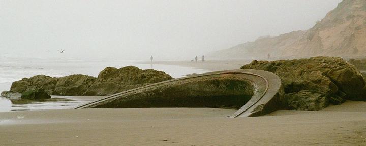 A concrete structure resembling a sunken ship is partially buried in the sand on a foggy beach with people and rocks in the distance.