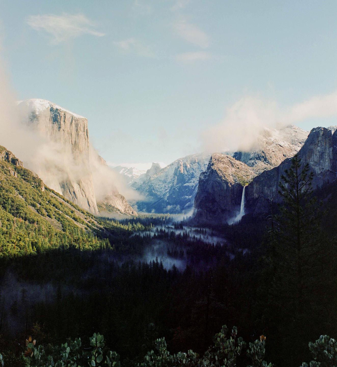 A valley with a waterfall and a forest is surrounded by mountains partially covered in clouds.