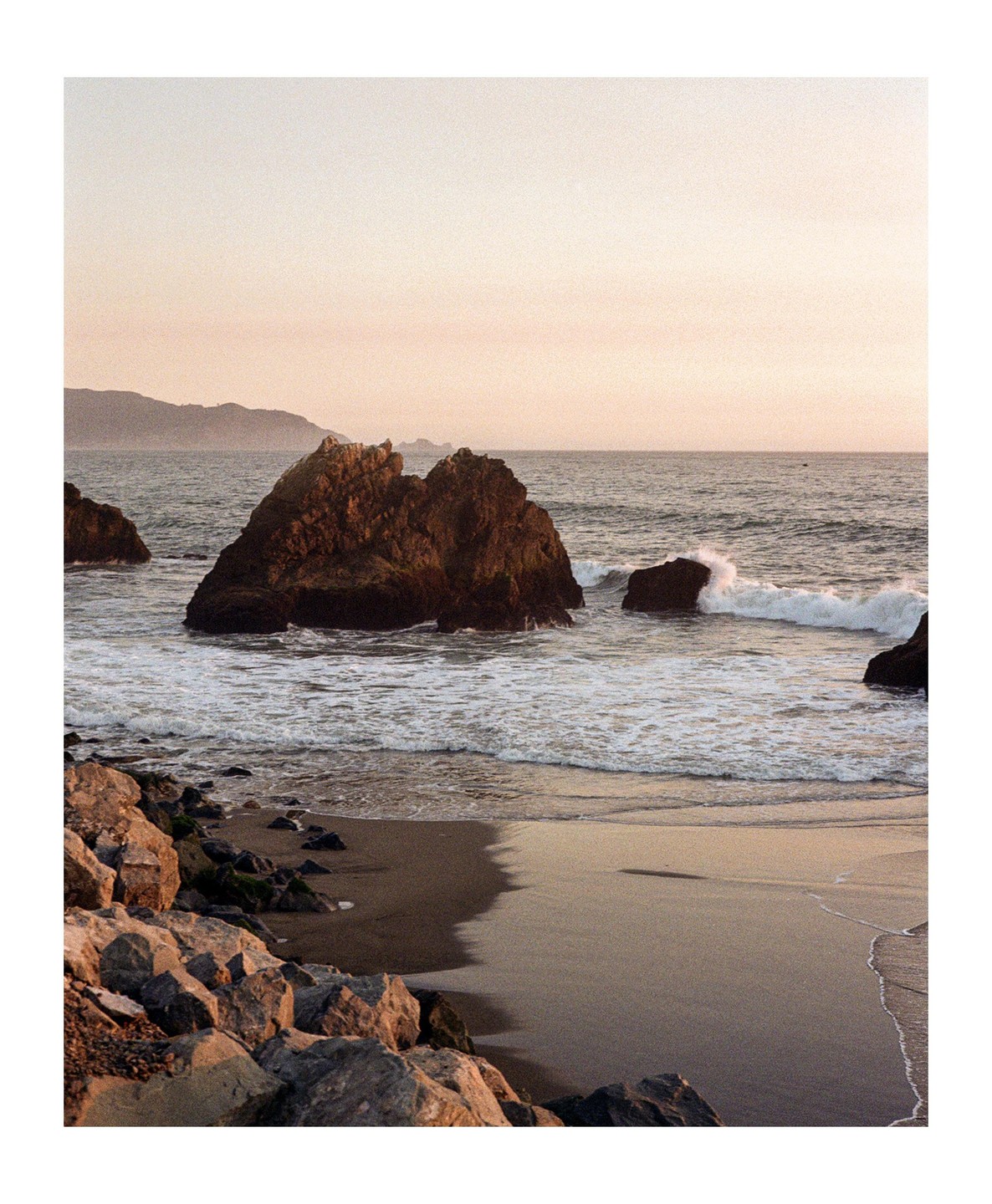 Waves crash against large rocks on a sandy beach with hills in the distance under a pale sky.
