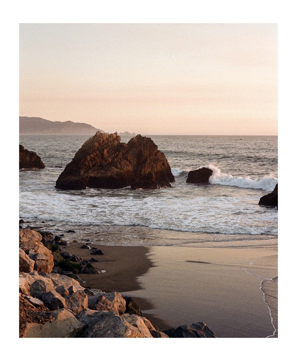 Waves crash against large rocks on a sandy beach with hills in the distance under a pale sky.