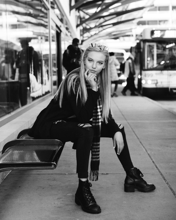 A woman with long hair and sunglasses on her head sits on a bench at a bus stop, looking at the camera.