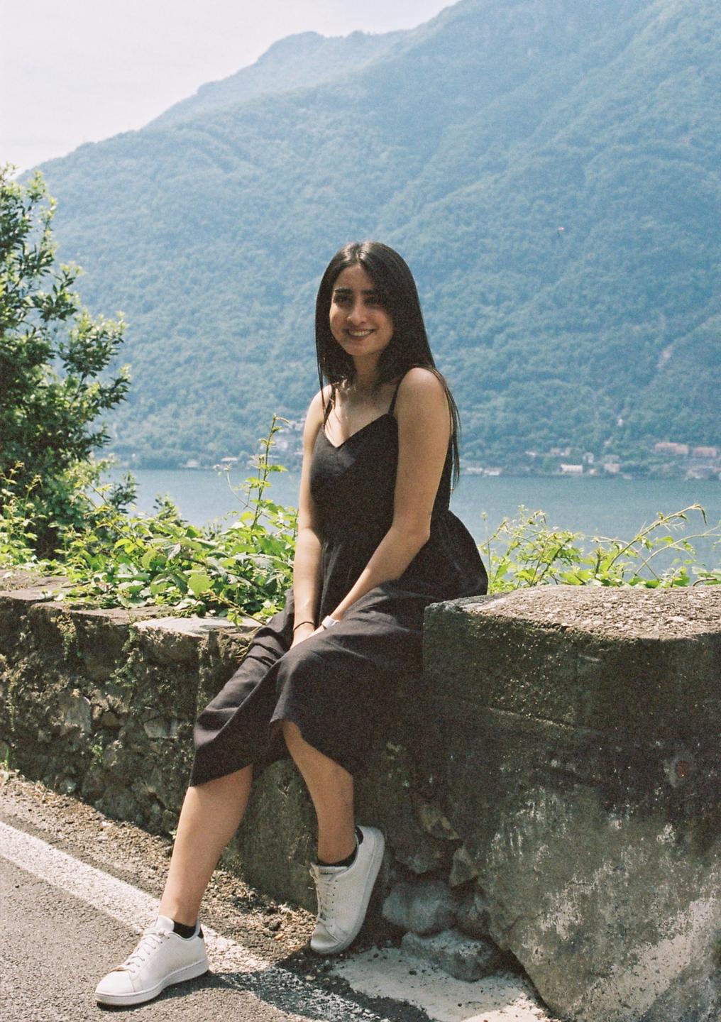 A woman in a black dress sits on a stone wall overlooking a lake and mountains.