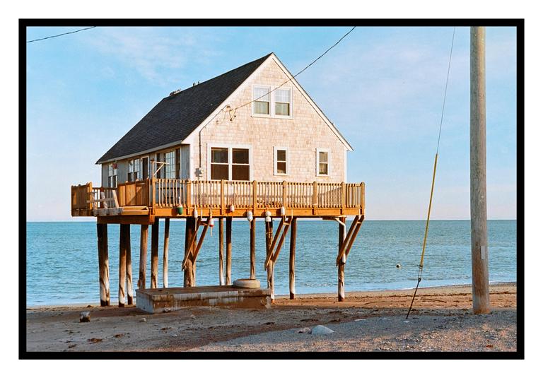 A house on stilts stands on a sandy beach next to the ocean under a clear sky.