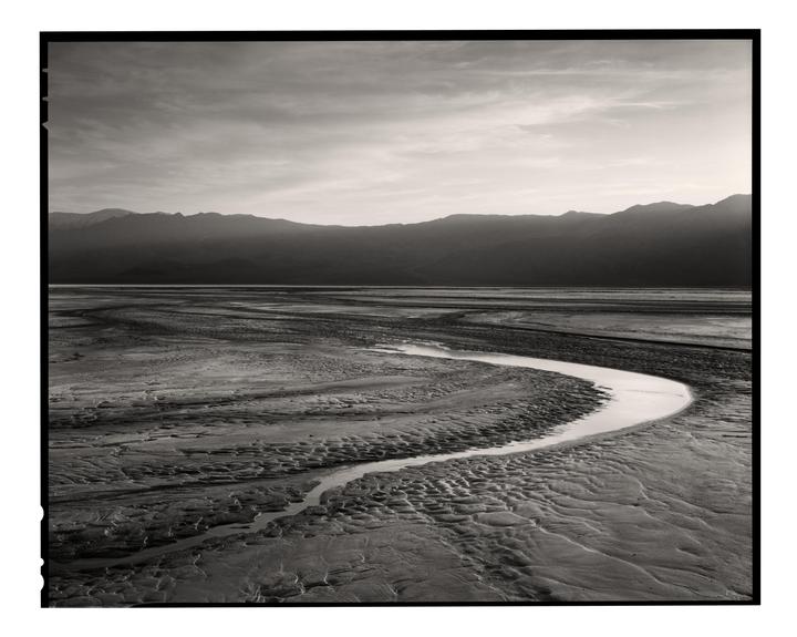 A winding stream flows through a textured desert landscape with mountains in the background under a cloudy sky.