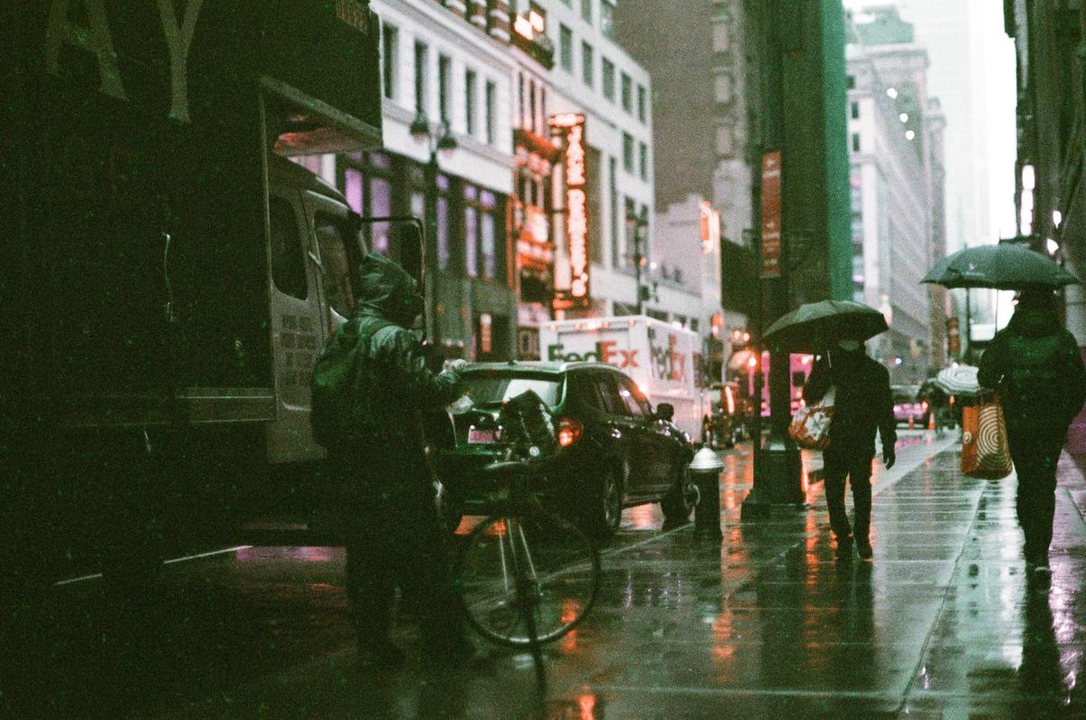 A person in a hooded jacket stands next to a bicycle and a parked car on a rainy street at night, while two other people walk with umbrellas in the background.