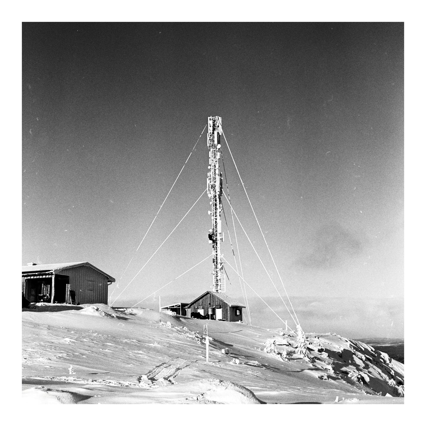 A tall communication tower stands on a snow-covered mountain with two small buildings nearby.