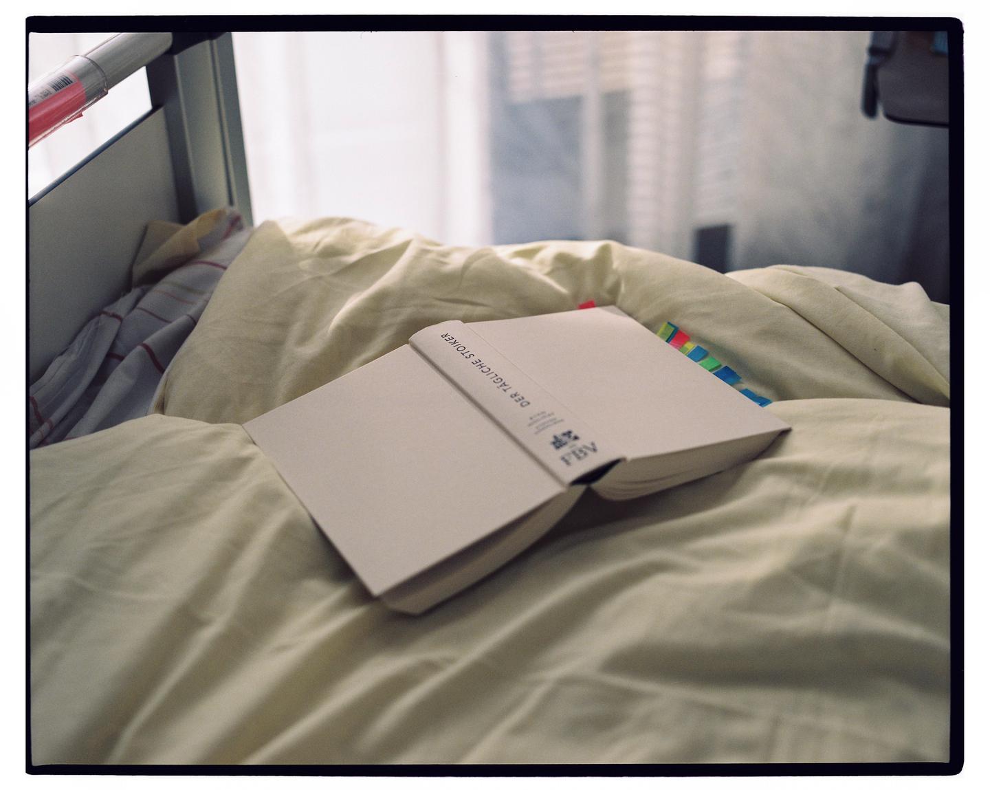 An open book with colorful page markers rests on a rumpled bed with light-colored sheets.