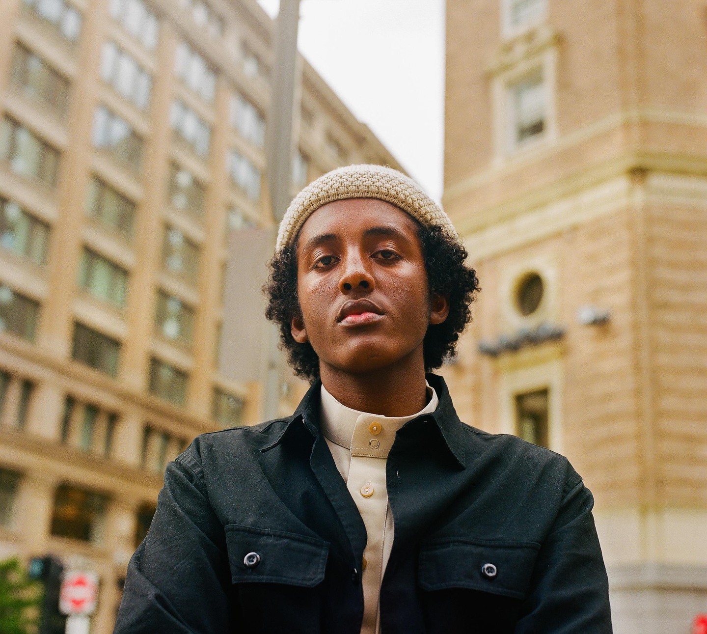 A young man with curly hair and a knit cap looks directly at the viewer, with buildings in the background.