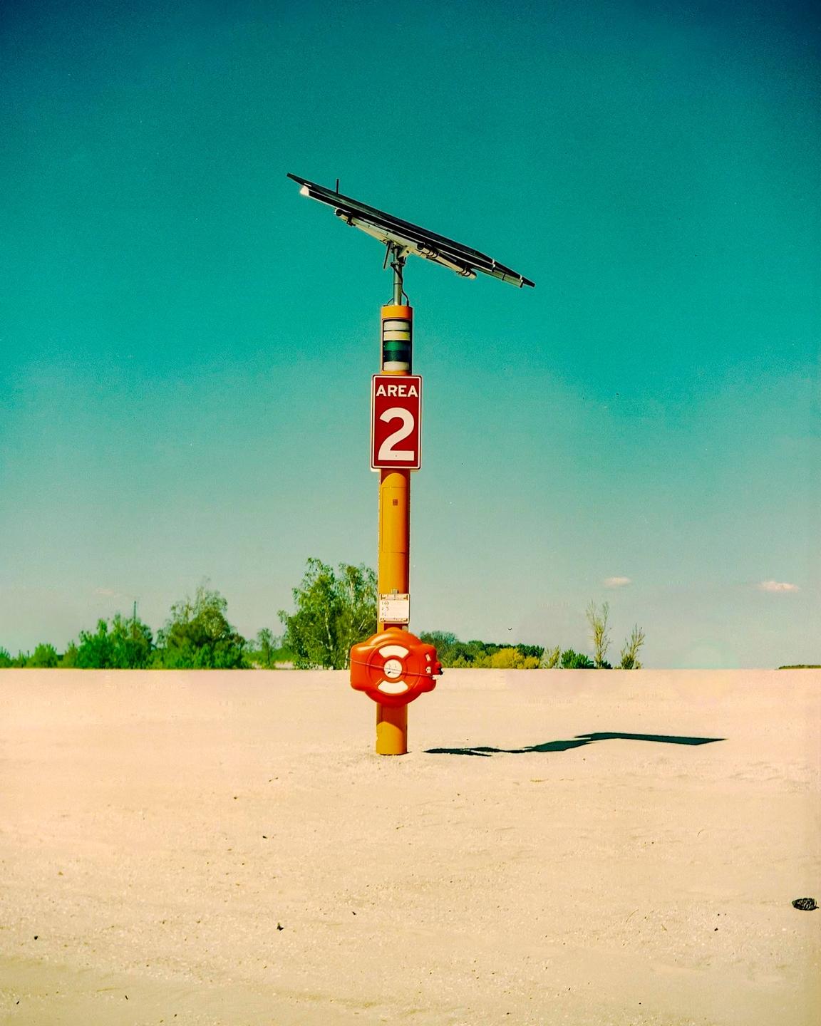 A yellow pole with a solar panel and an "AREA 2" sign stands on a sandy beach under a clear blue sky.