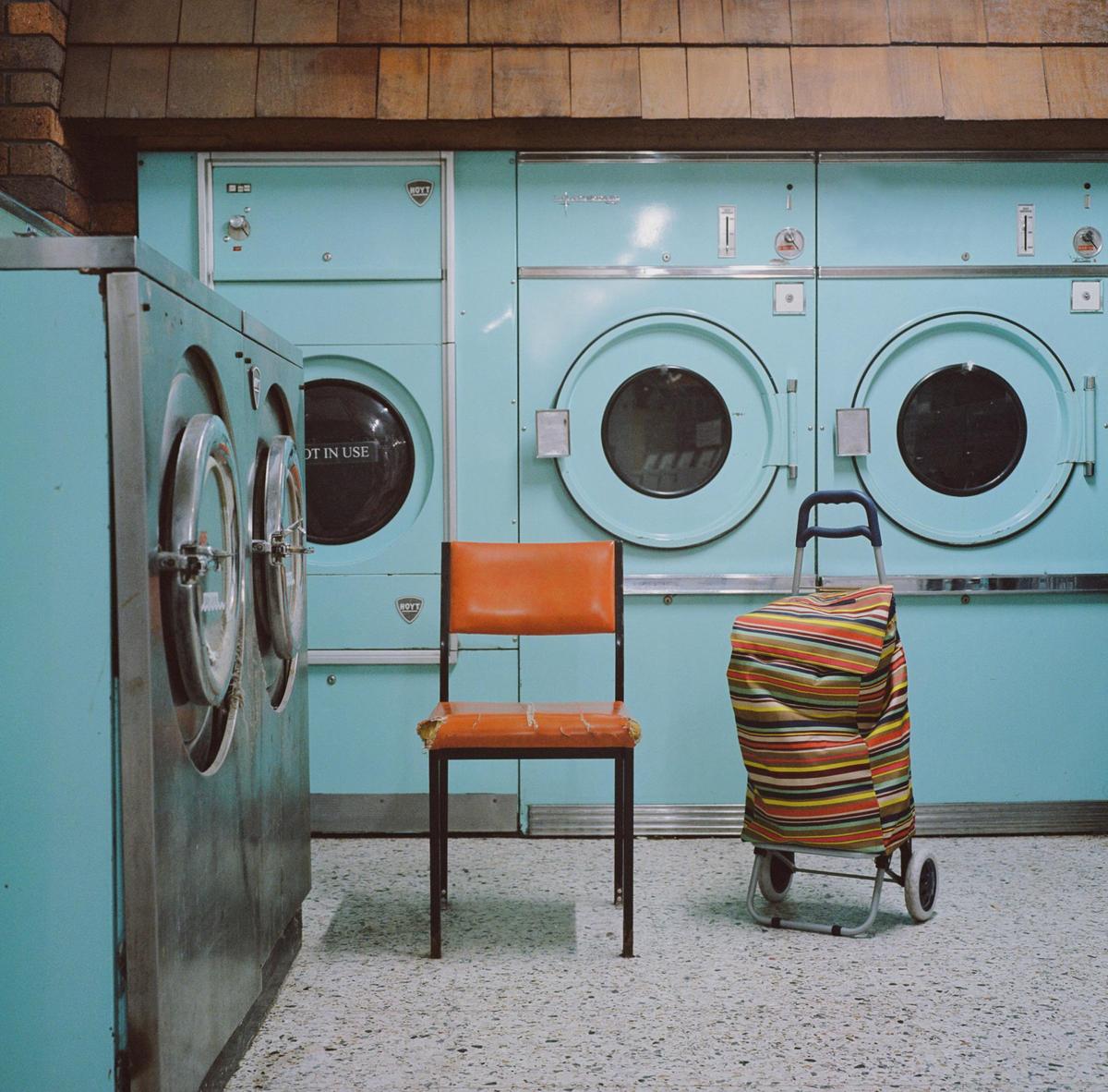 An orange chair and a striped laundry cart sit in front of a row of light blue washing machines.