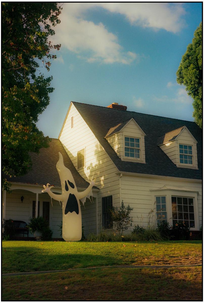 A large inflatable ghost stands in the front yard of a white house with a dark roof.