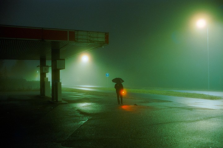 A person holding an umbrella and a red light stands under a gas station canopy on a foggy night.