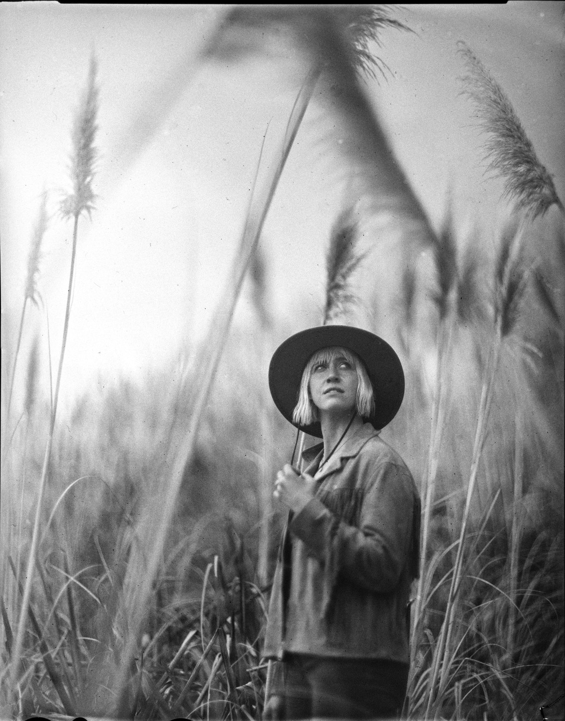 A woman in a hat stands among tall grasses, looking up.