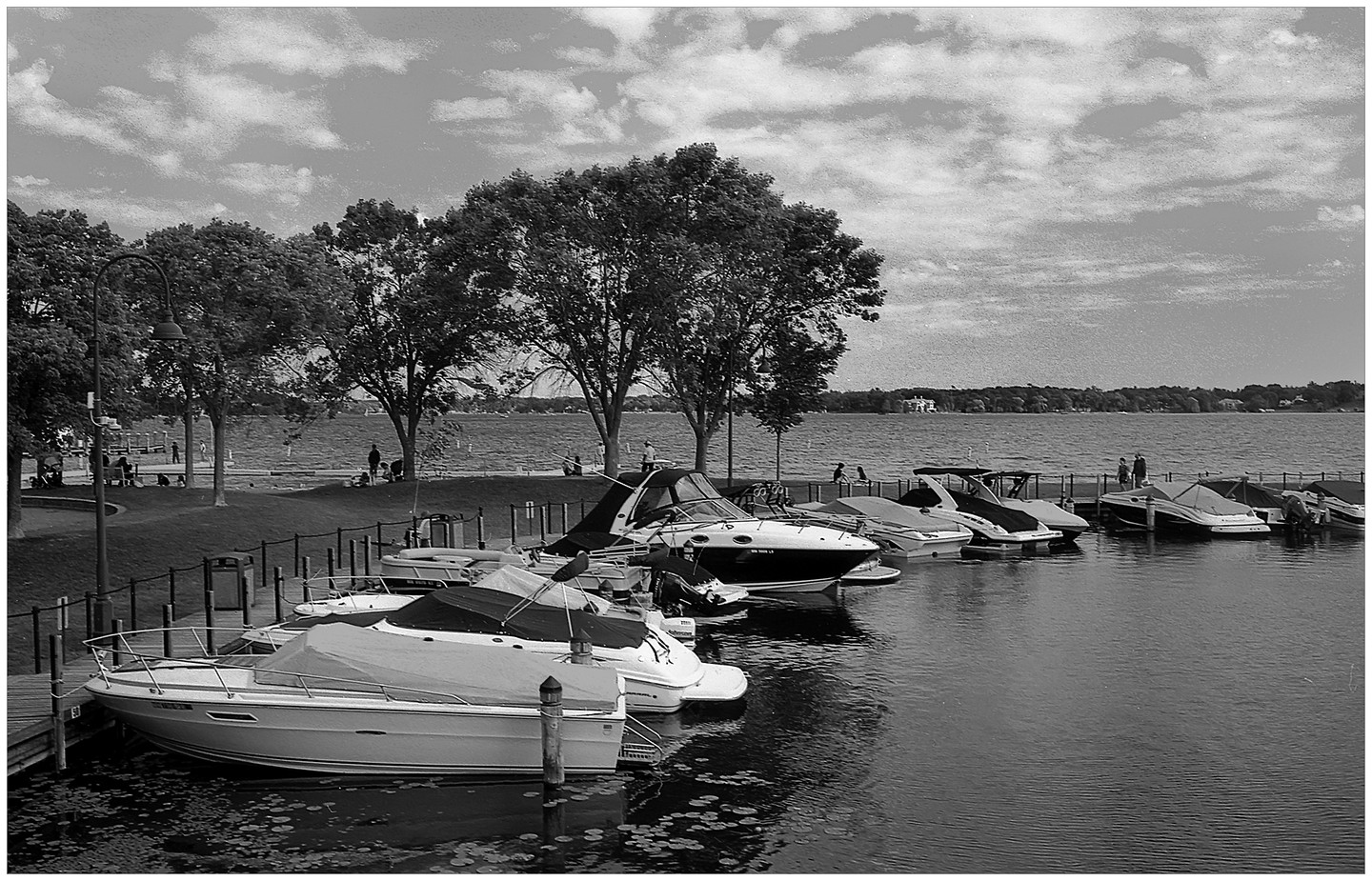 Boats are docked along a lakefront with trees and people in the background under a cloudy sky.