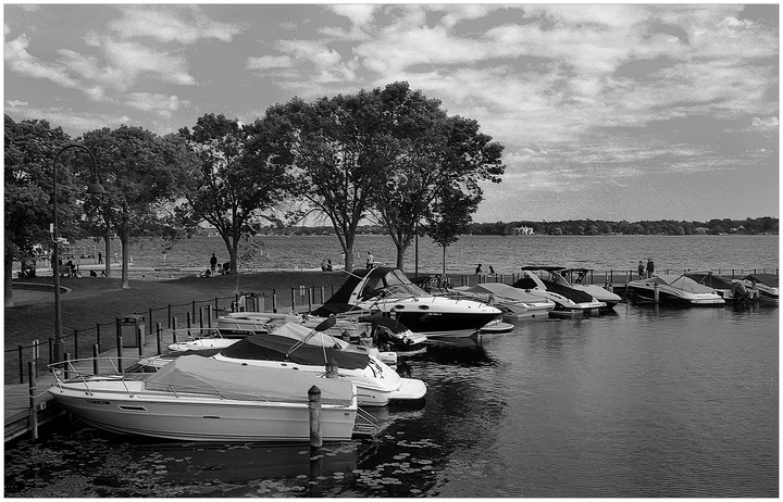 Boats are docked along a lakefront with trees and people in the background under a cloudy sky.