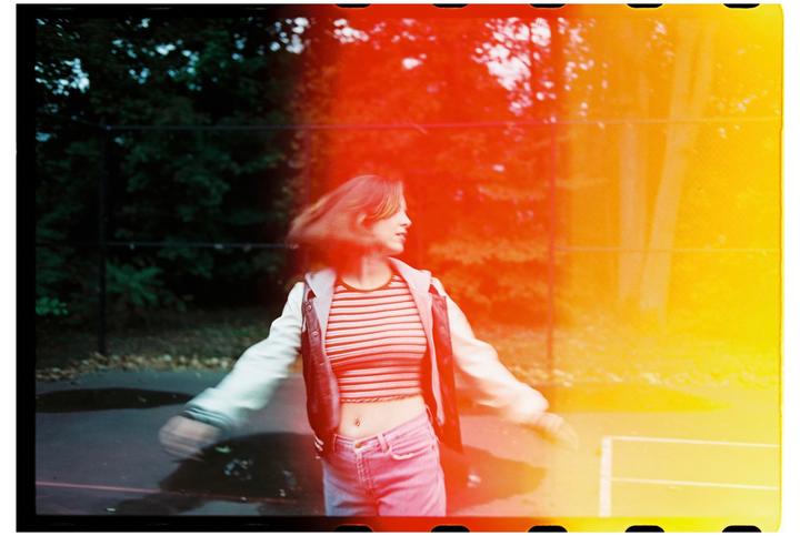 A woman with her hair in motion stands on a court with trees in the background, with a strong red and yellow light leak across the image.
