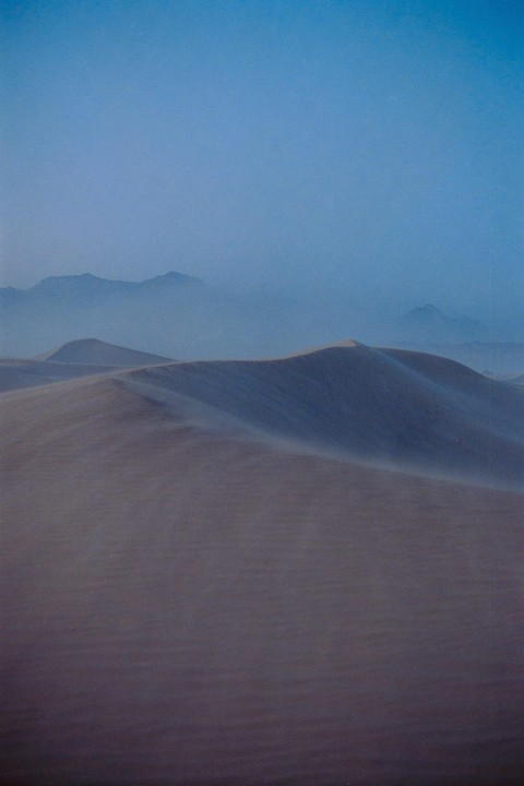 Sand dunes are visible with mountains in the distance under a hazy blue sky.
