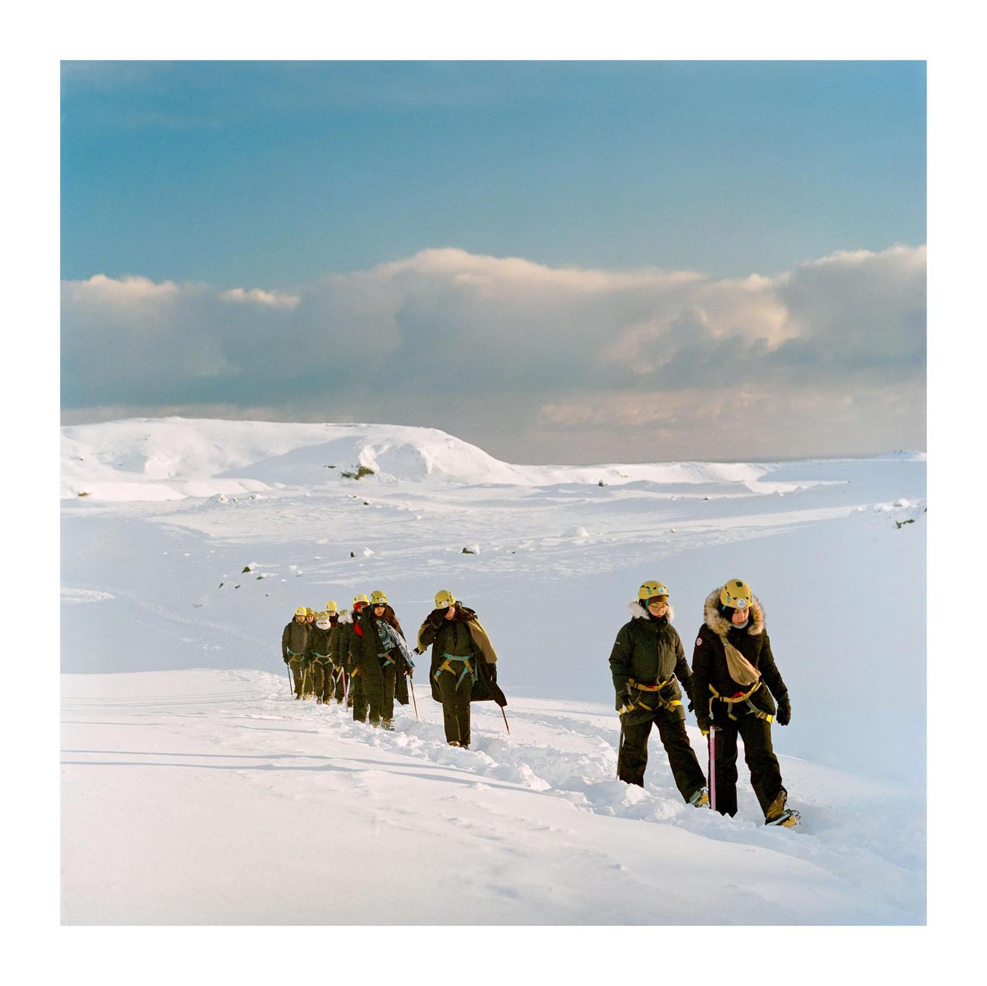 A group of people wearing winter gear and helmets walk in a line through a snow-covered landscape under a partly cloudy sky.