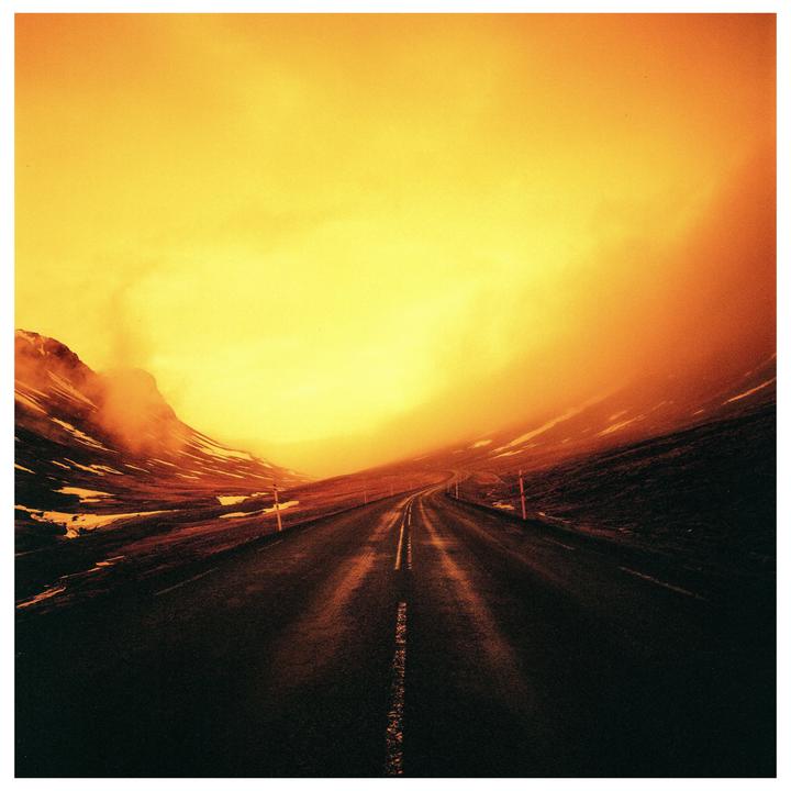 A road stretches into the distance through a mountainous landscape under a bright orange sky.
