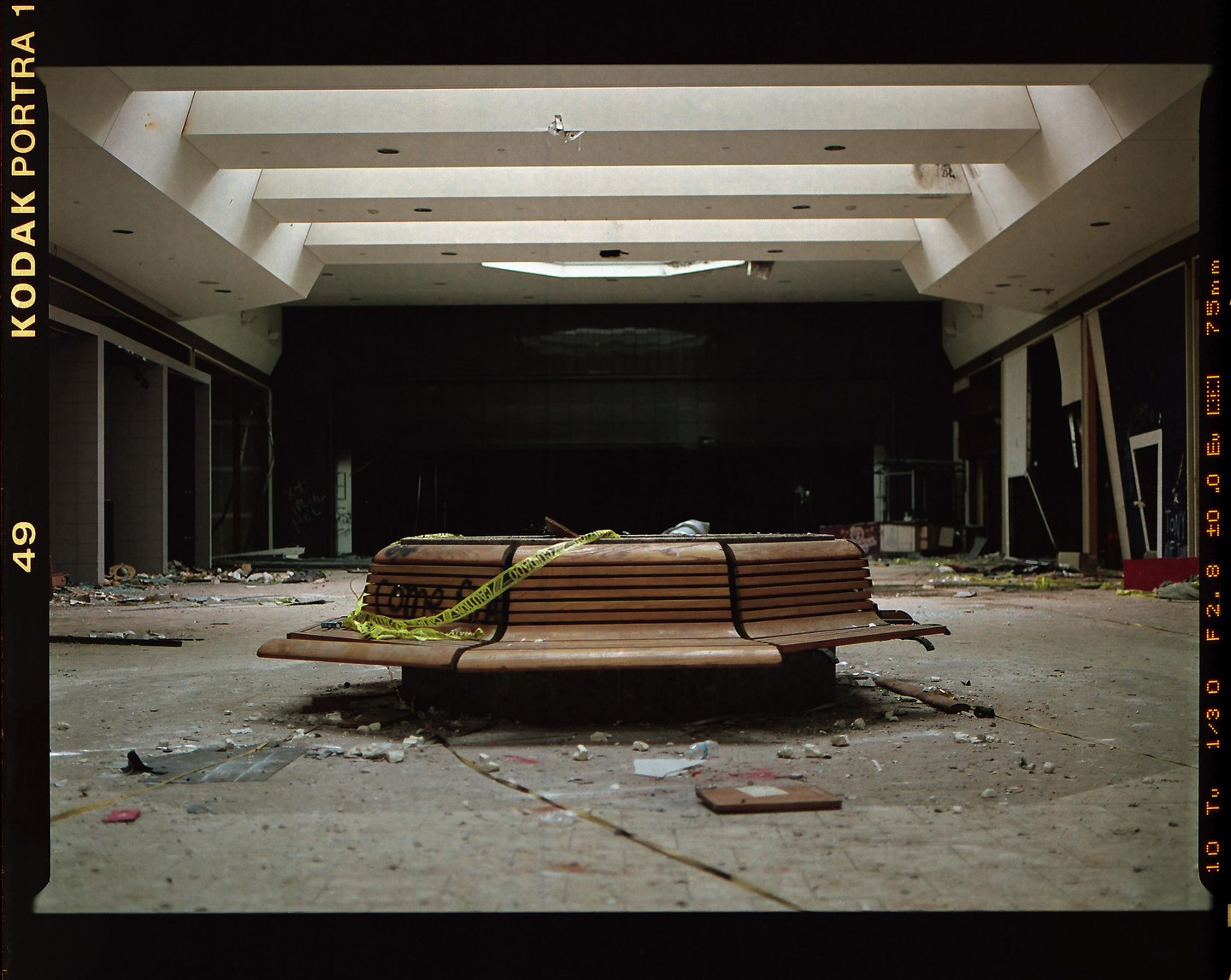 A wooden bench with yellow caution tape sits in the middle of a derelict indoor space with debris scattered on the floor.
