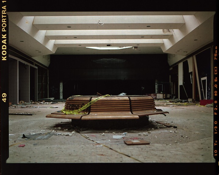 A wooden bench with yellow caution tape sits in the middle of a derelict indoor space with debris scattered on the floor.