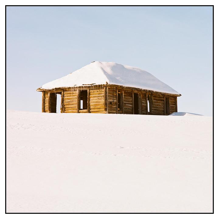A wooden cabin with a snow-covered roof stands in a field of snow under a clear sky.