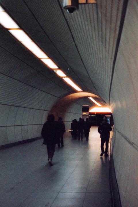 People walk through a subway tunnel with bright lights on the ceiling.