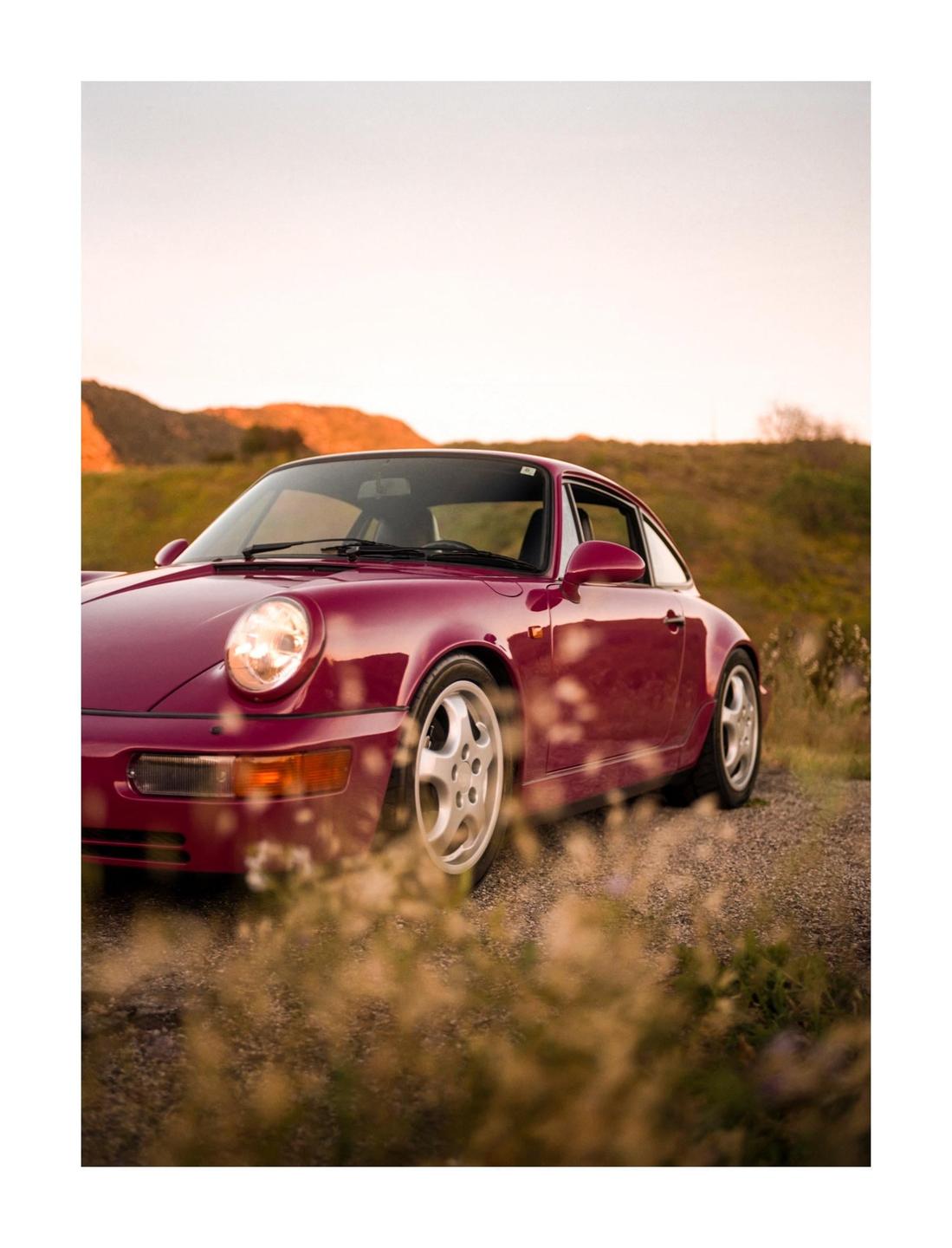 A pink car is parked on a gravel road with hills in the background.
