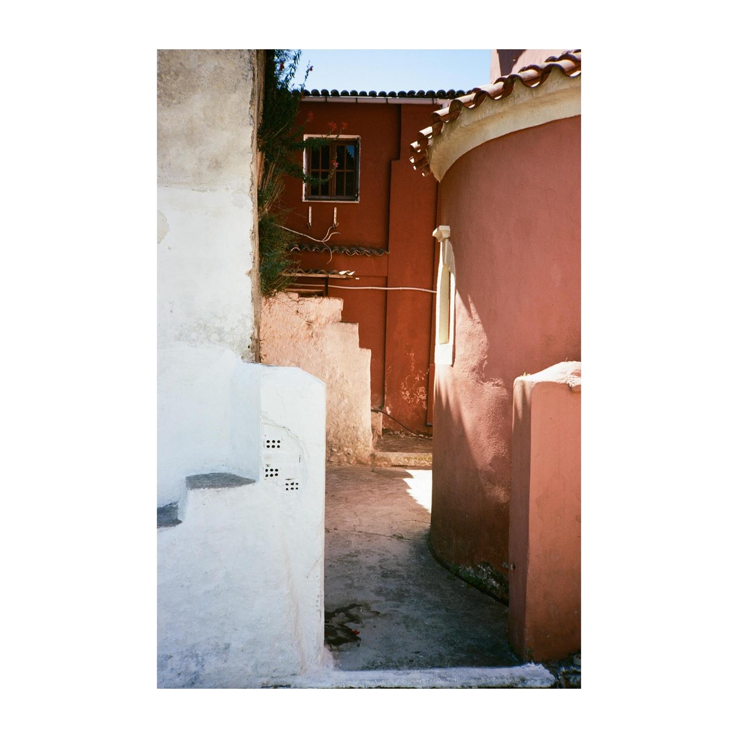 A narrow alleyway between buildings with white and red walls leads to a sunlit path.