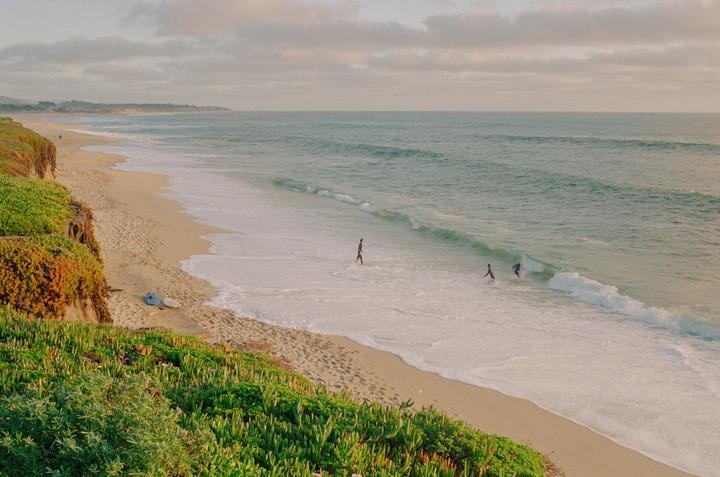 Two people are in the ocean near a sandy beach with green vegetation on a cliff.