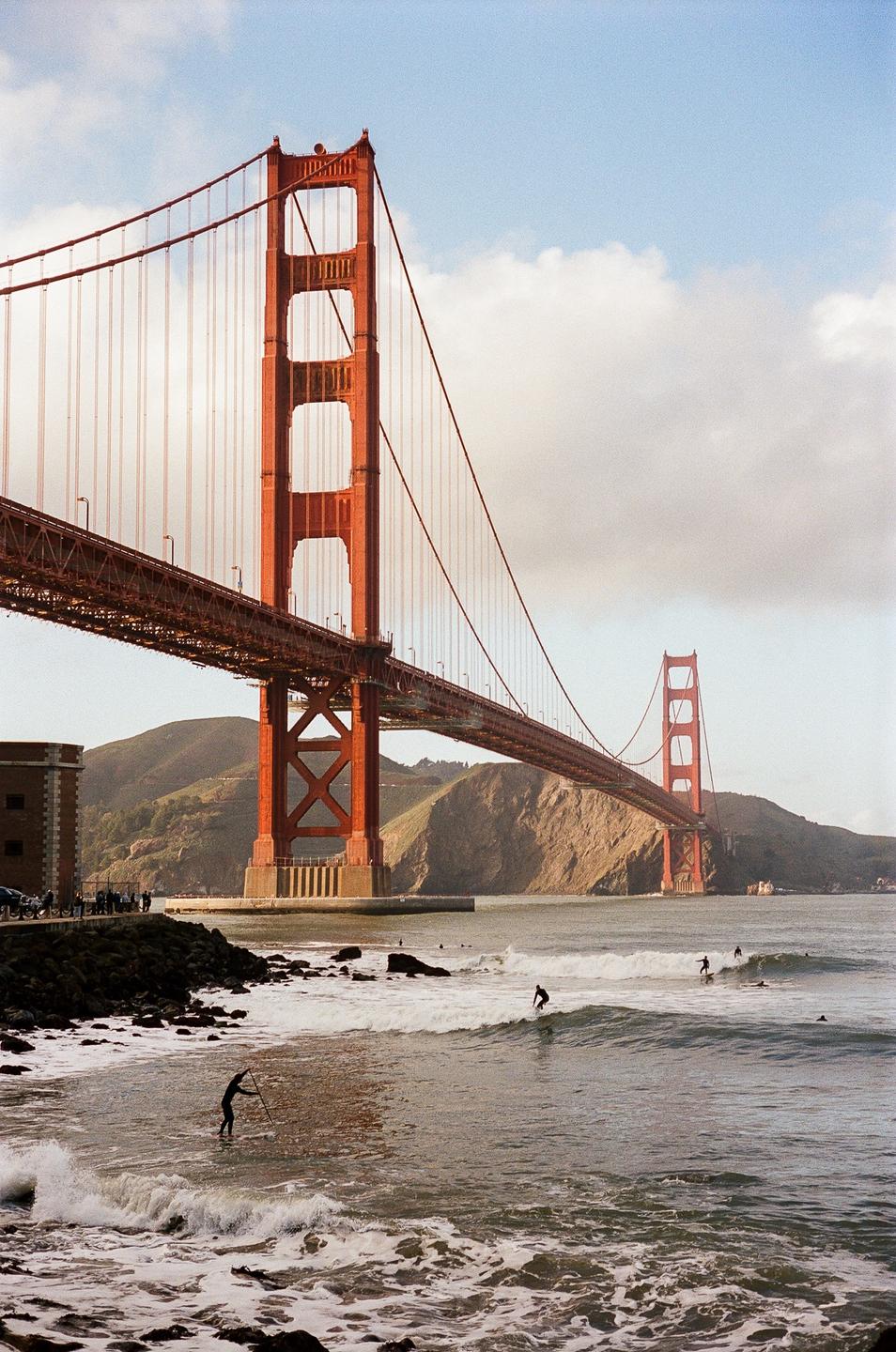 The Golden Gate Bridge spans a body of water where surfers ride waves near the shore.