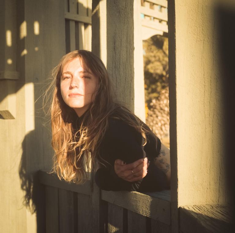 A woman with long hair leans on a wooden railing, with sunlight casting shadows on her face and the wall behind her.