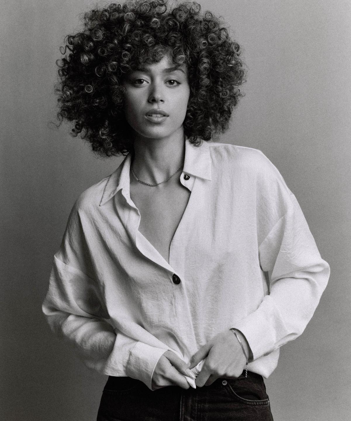 A woman with curly hair wears a white shirt and dark pants while looking at the viewer.