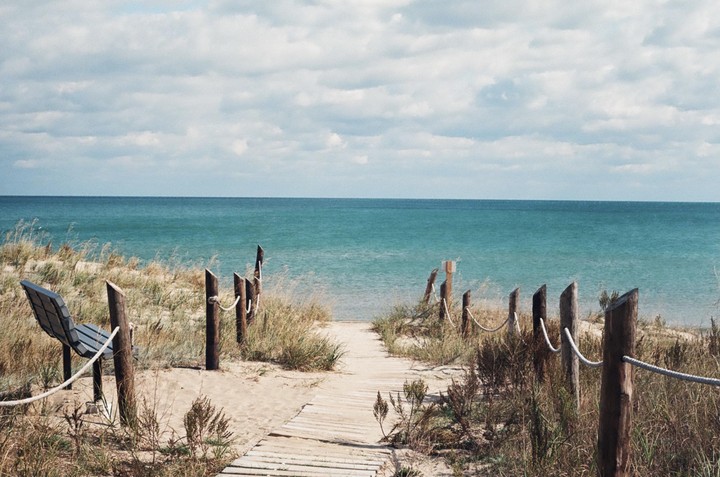 A wooden boardwalk leads through sandy dunes towards a vast blue ocean under a cloudy sky.