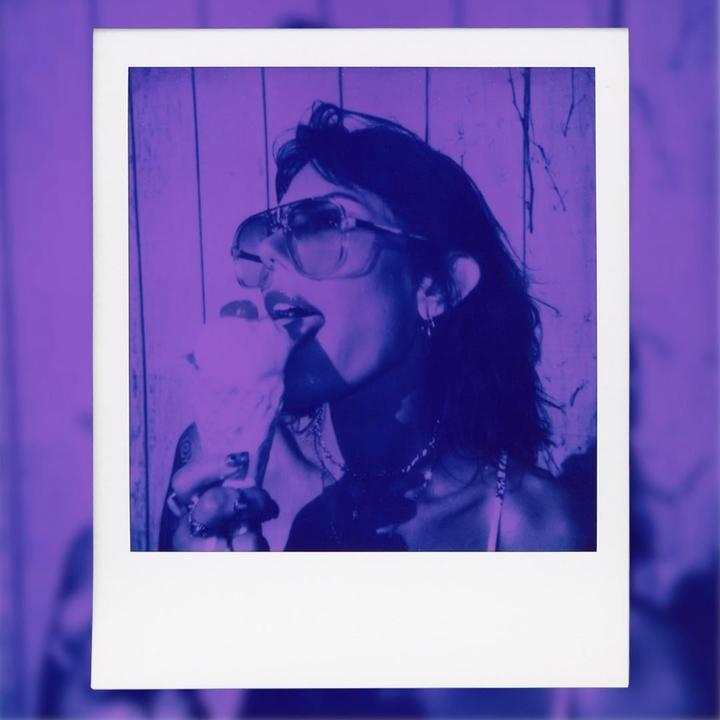A woman wearing sunglasses licks an ice cream cone in a purple-toned image.