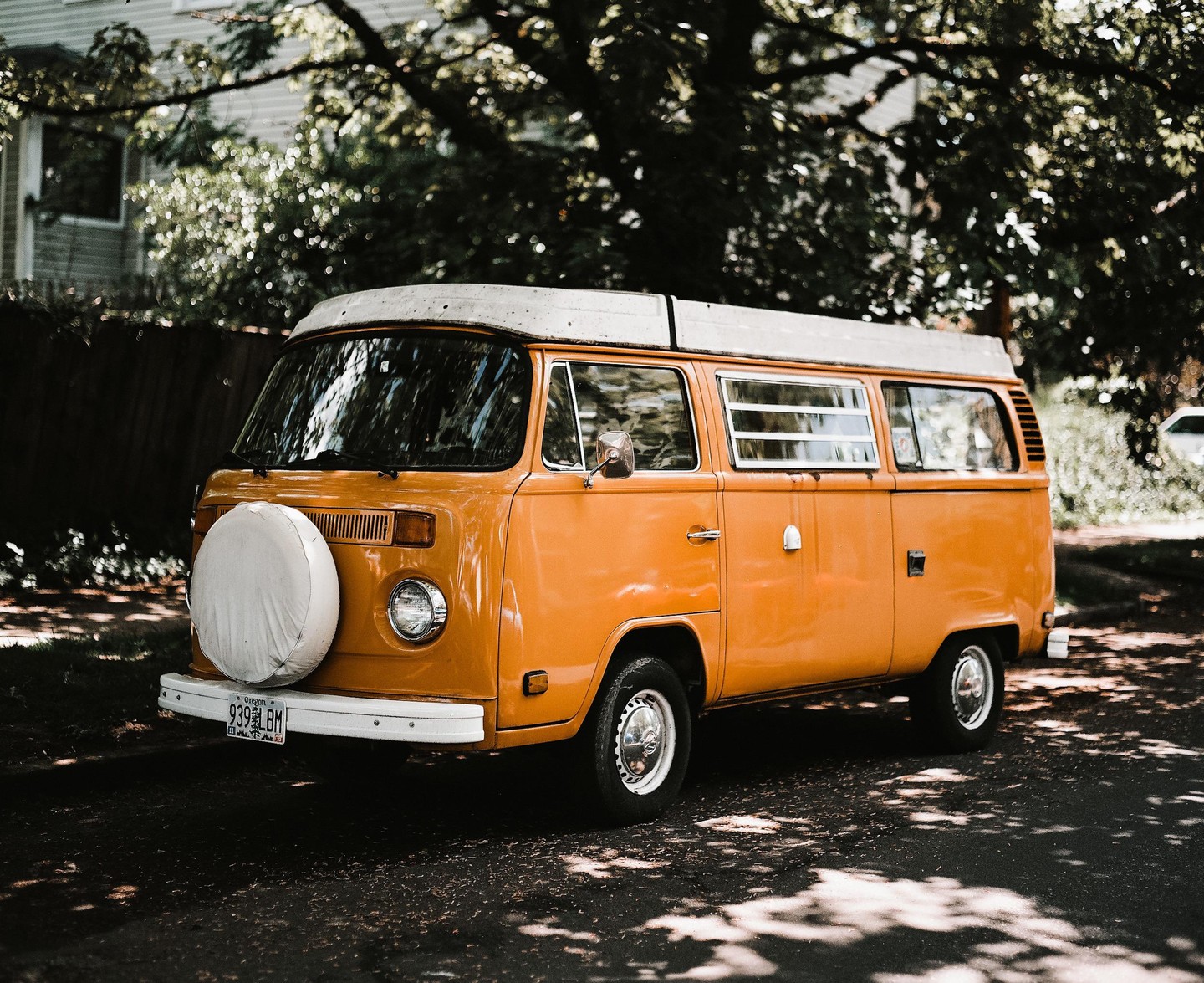 An orange van with a white roof and a spare tire on the front is parked on a shaded street.