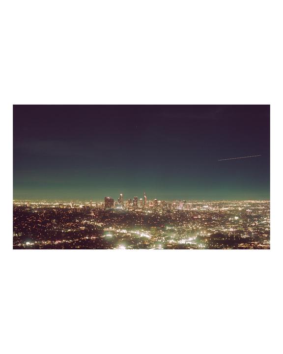 A panoramic view of the Los Angeles skyline at night, with city lights stretching into the distance and a faint airplane contrail visible in the dark sky.