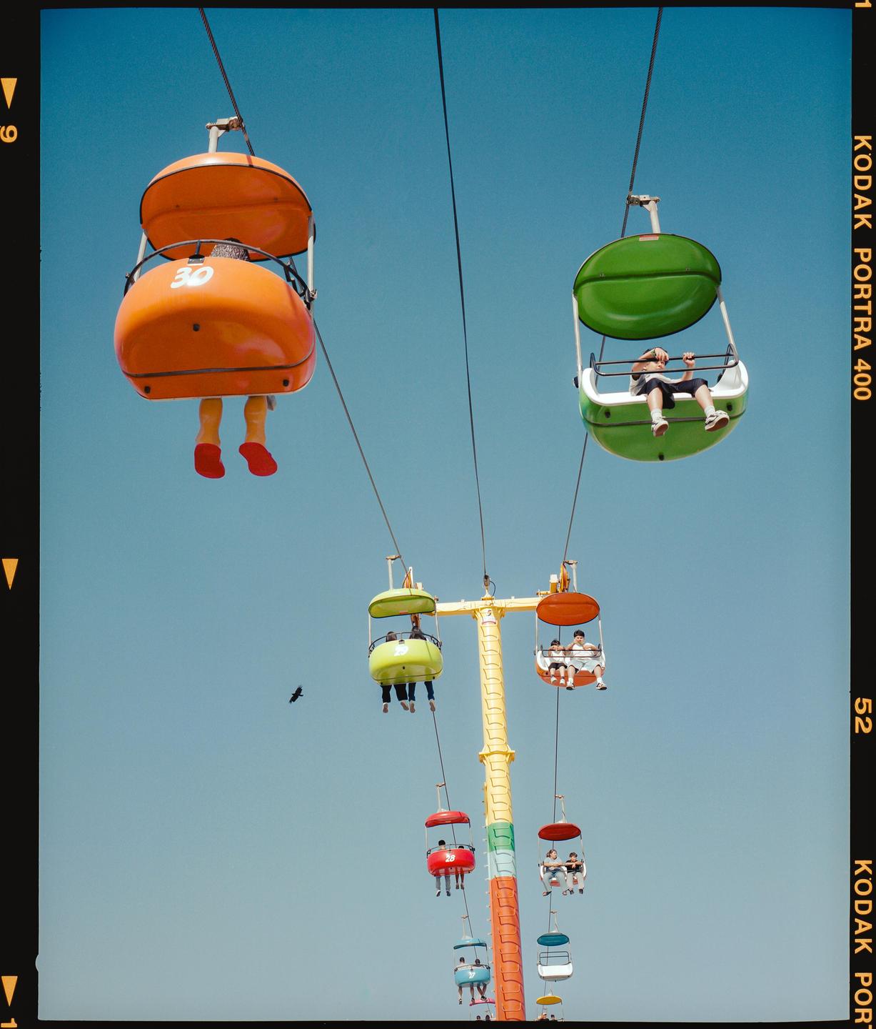 People ride in colorful gondolas on a sky ride attraction against a clear blue sky.