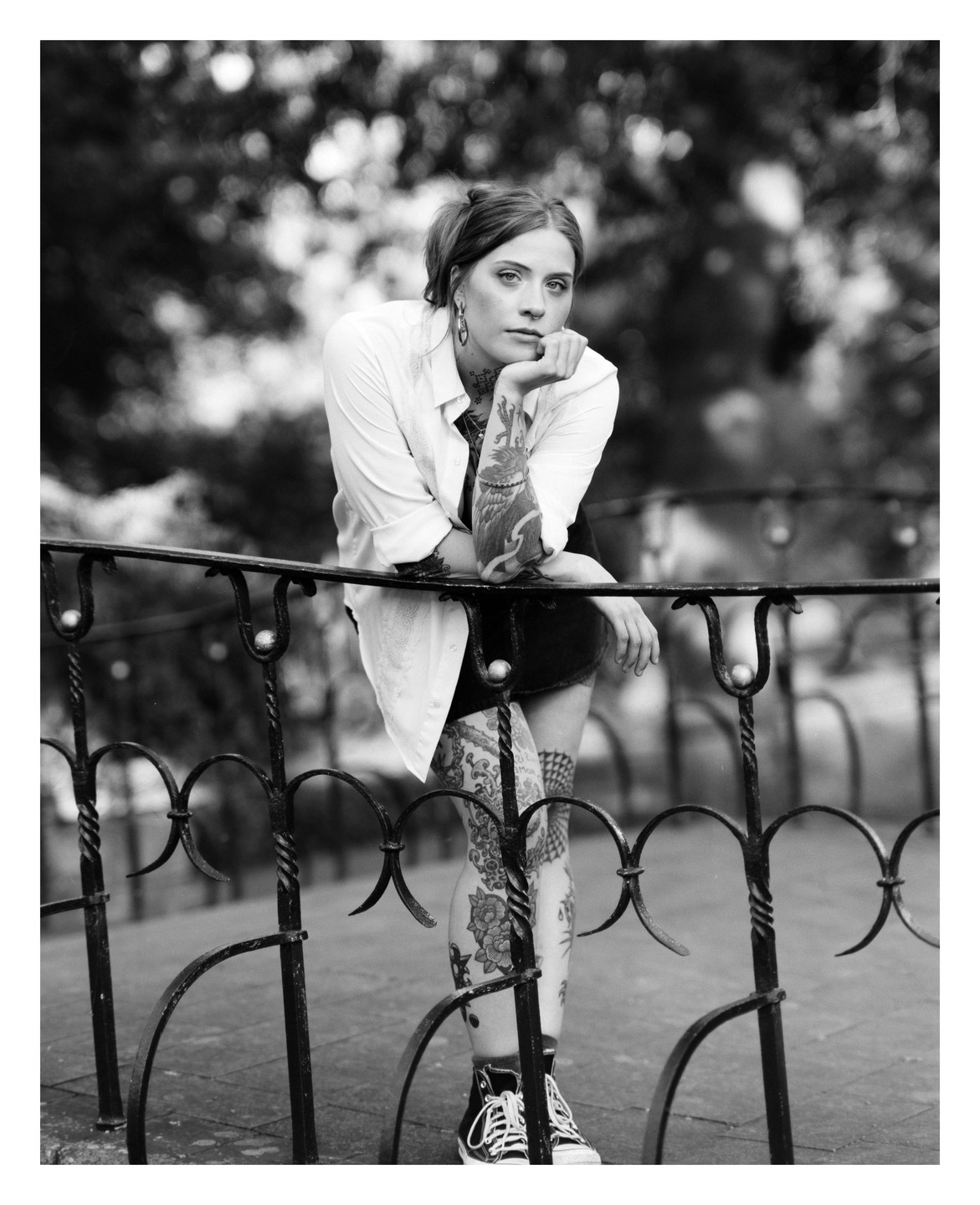 A woman with tattoos on her arms and legs leans on a metal fence.