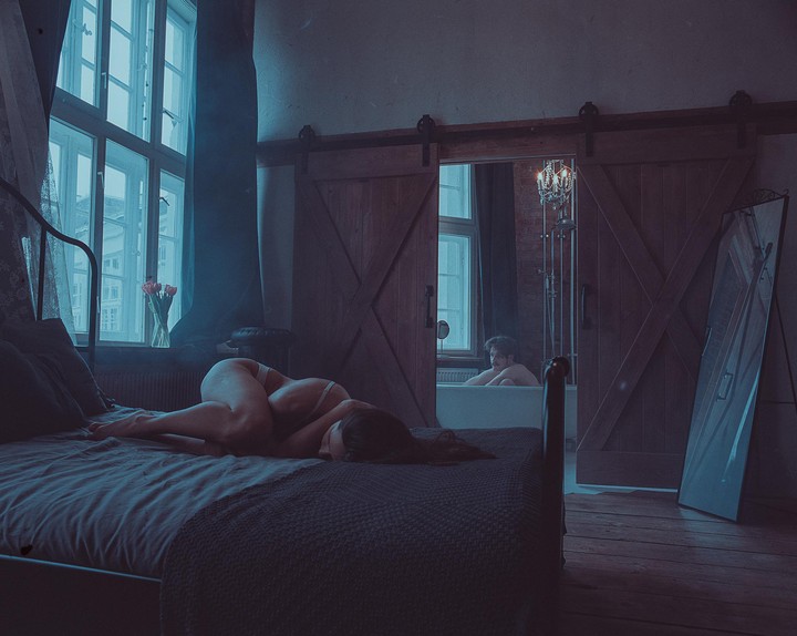 A woman lies on a bed in a dimly lit room while a man sits in a bathtub visible through an open doorway.