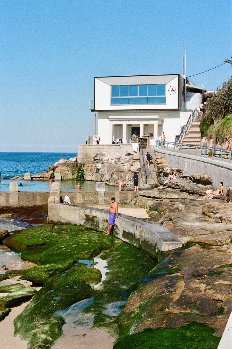 A white building with a clock on its facade overlooks a rocky beach with people swimming and sunbathing.