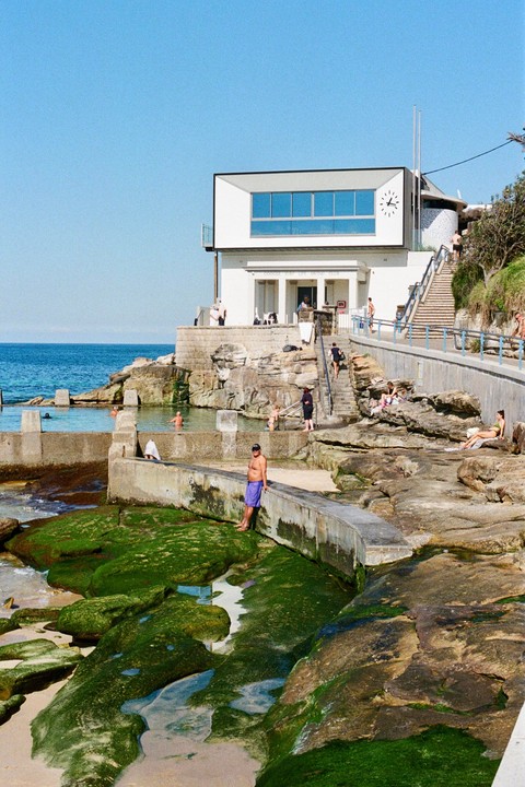 A white building with a clock on its facade overlooks a rocky beach with people swimming and sunbathing.