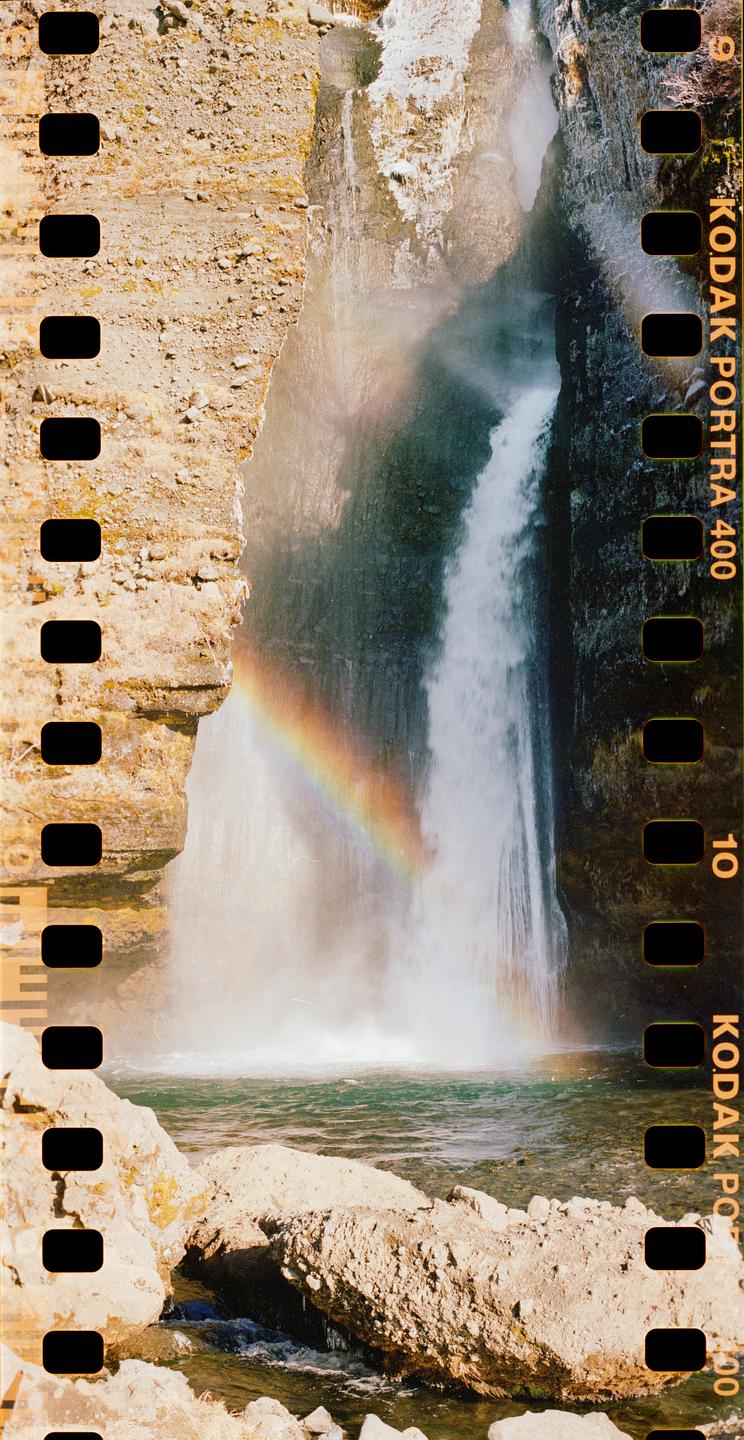 A rainbow is visible in the mist at the base of a waterfall cascading between rocky cliffs.