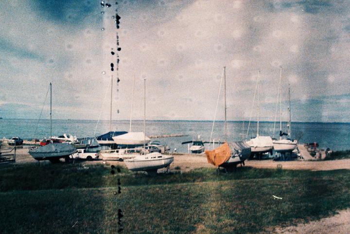 Several sailboats are dry-docked on a grassy and dirt area next to a body of water under a cloudy sky.