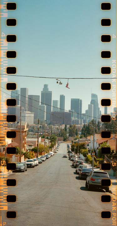 A street with parked cars leads to a city skyline with shoes hanging from a power line.