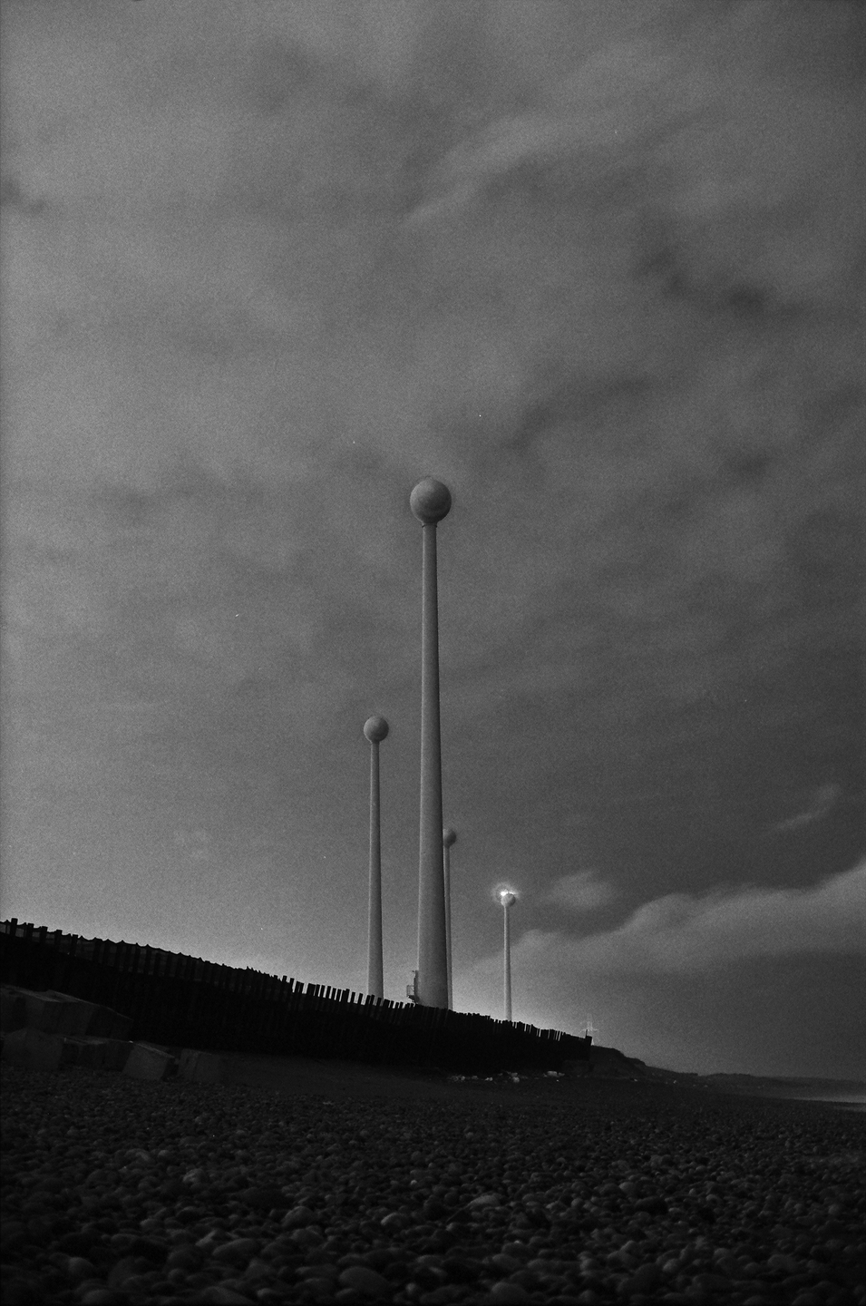 A black and white image shows tall, spherical streetlights on a pebble beach under a cloudy sky.