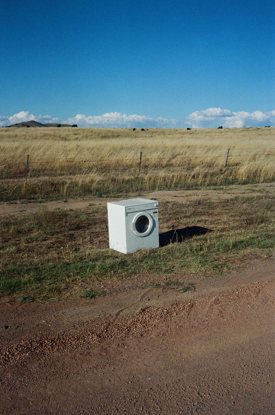 A washing machine sits abandoned on a dirt road next to a field of dry grass under a clear blue sky.