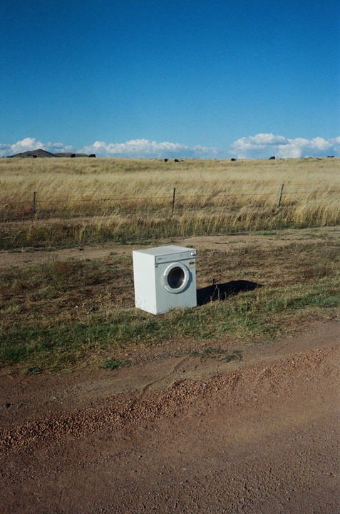 A washing machine sits abandoned on a dirt road next to a field of dry grass under a clear blue sky.