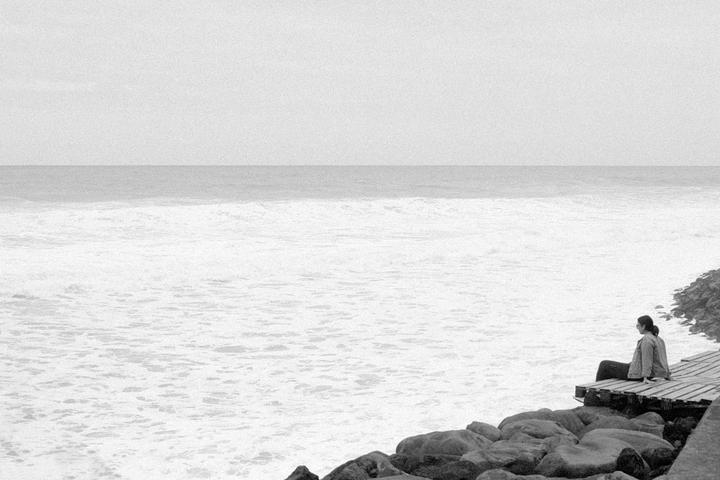 A woman sits on a wooden platform overlooking a vast ocean with foamy waves.